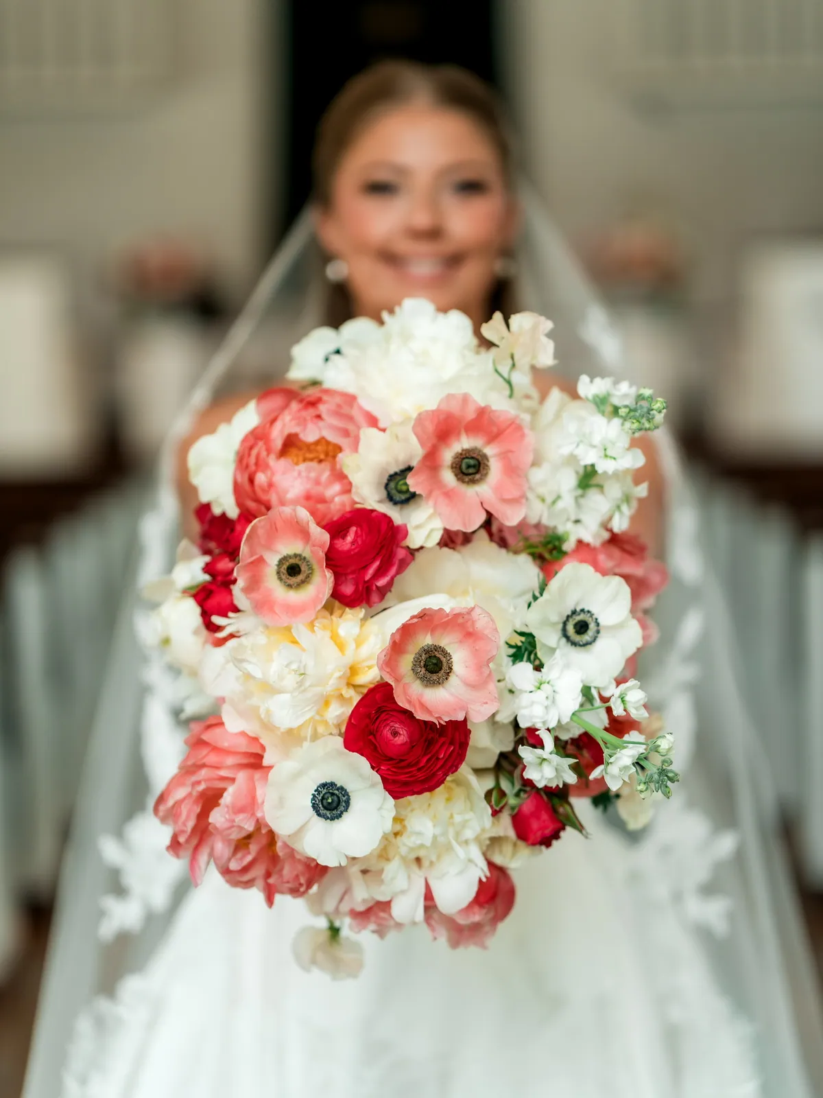 Coral peony, white anemone and red ranunculus bridal bouquet held toward the camera in a chapel aisle