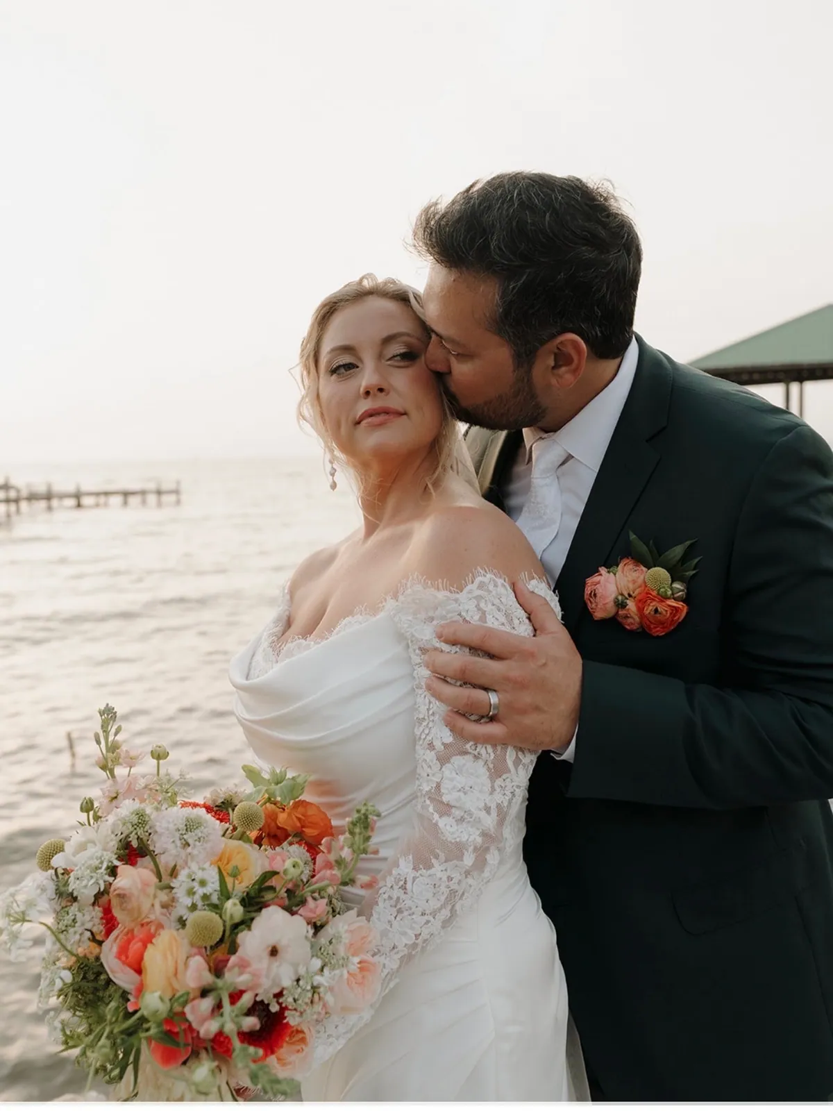 Bride and groom on a bay pier at dusk with a loose coral, peach and white bouquet and a matching boutonniere