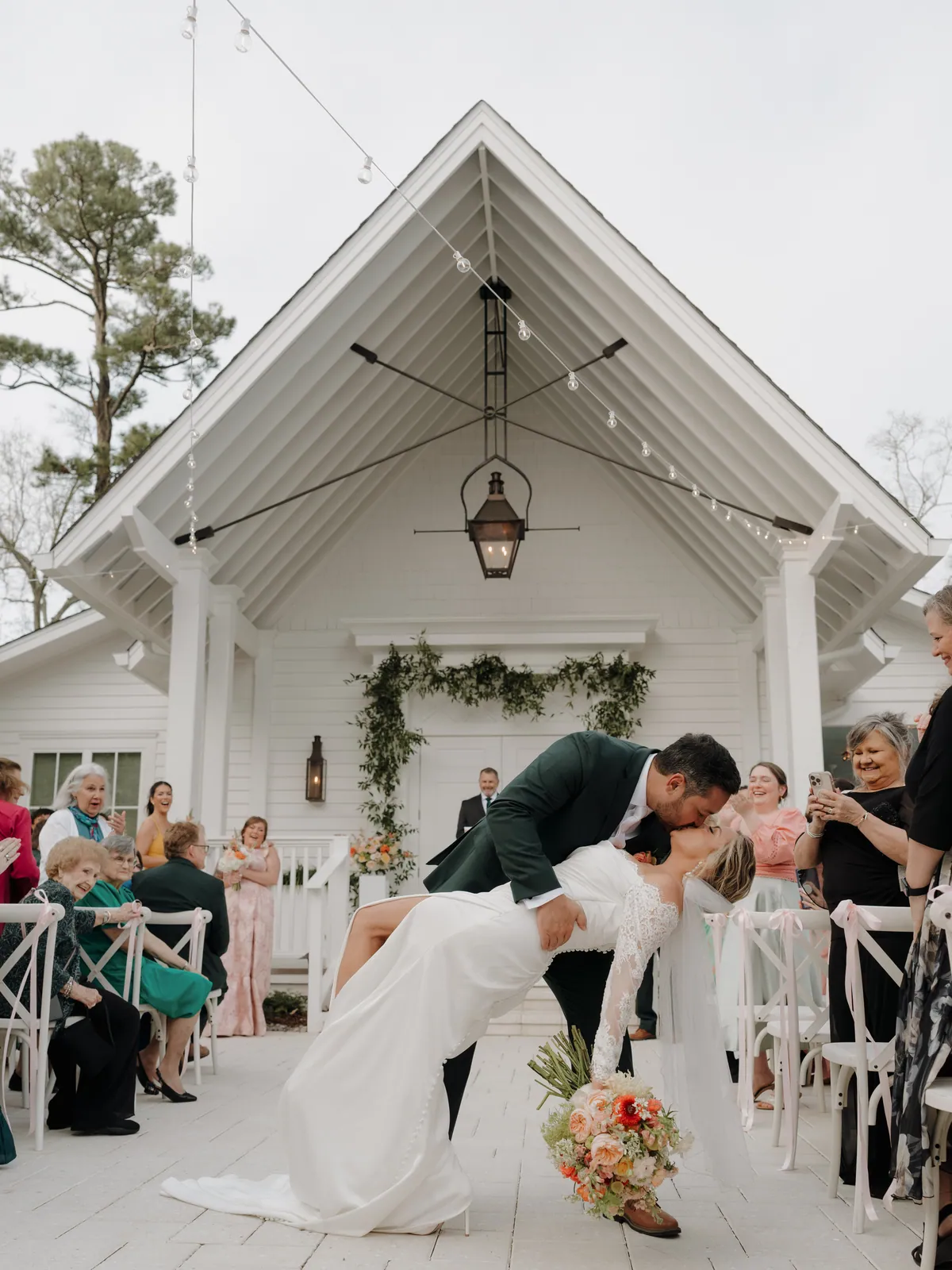Groom dipping his bride outside a white chapel hung with greenery, her peach and coral bouquet held out to the side